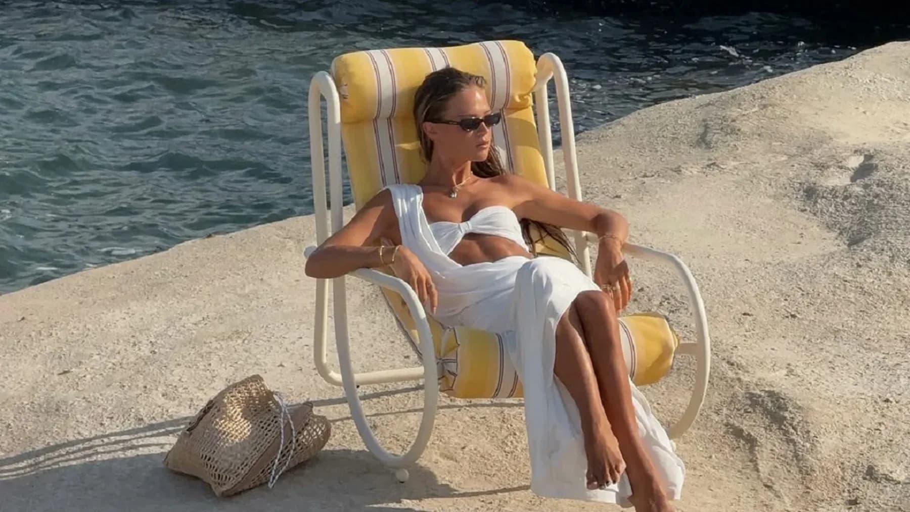 Žena u beloj letnjoj odeći i sa naočarima za sunce sedi u ležaljci pored mora, uz pletenu torbu na tlu.