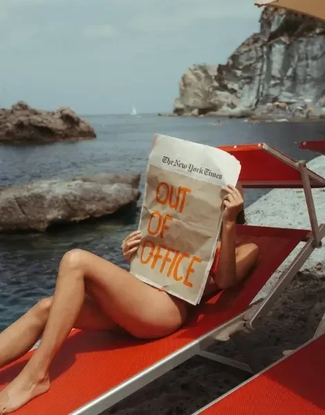Devojka leži na crvenoj ležaljci pored mora držeći novine sa natpisom Out of office
