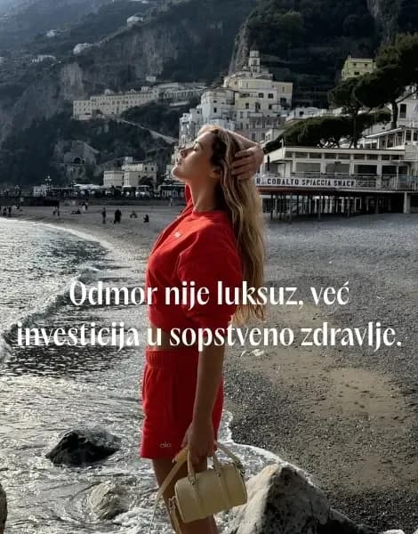 Devojka u crvenom kompletu stoji na obali ispred primorskog gradića na litici